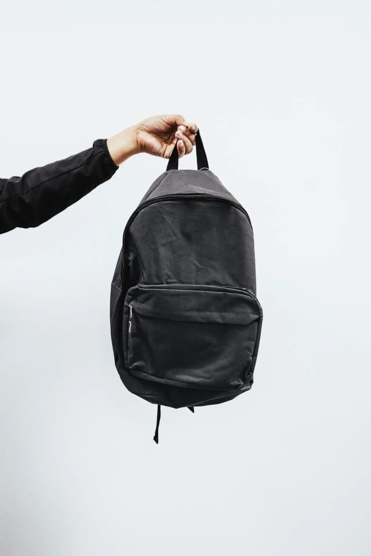 A simple black backpack being held by a hand against a minimal white background, emphasizing style and practicality.